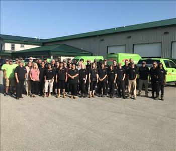 Large group of SERVPRO staff members standing in front of a tan warehouse with green trucks inside