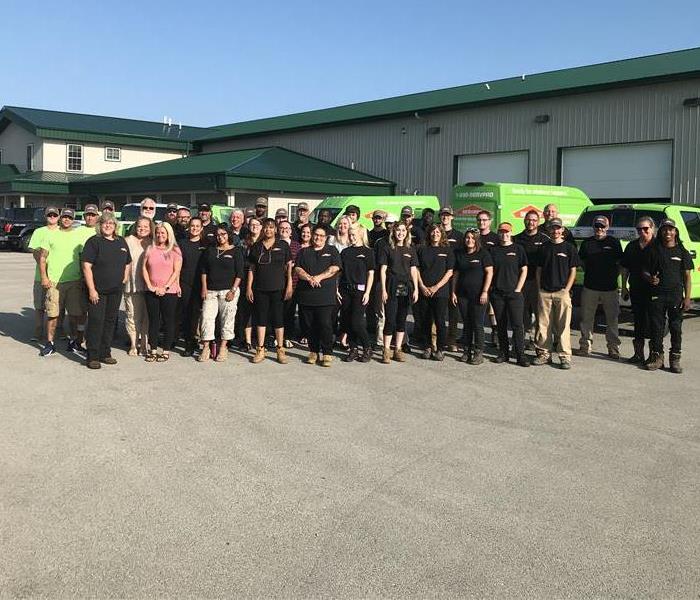 A large group of SERVPRO employees standing in front of a warehouse with green trucks inside
